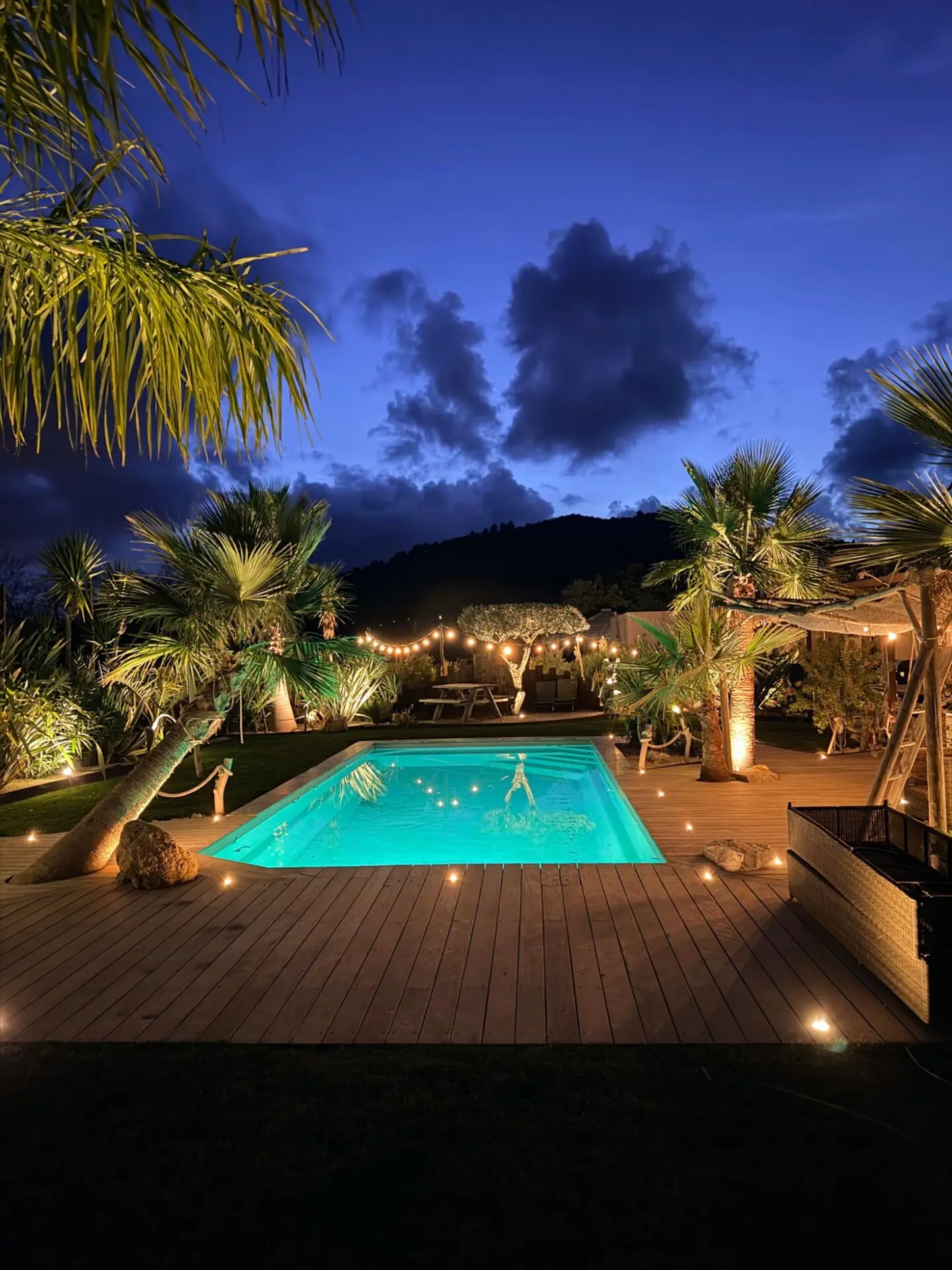 Piscine éclairée à la tombée de la nuit, terrasse en bois, palmiers et guirlandes